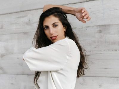 Catherine Coury with wit her right arm bent behind her head and her left arm crossing her body; wearing a white top against a gray wooden background