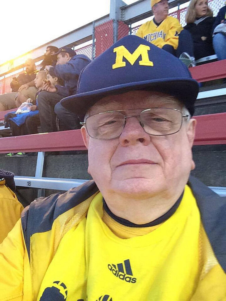 John Morgan wears Michigan attire, seated in the stands at an athletic event.