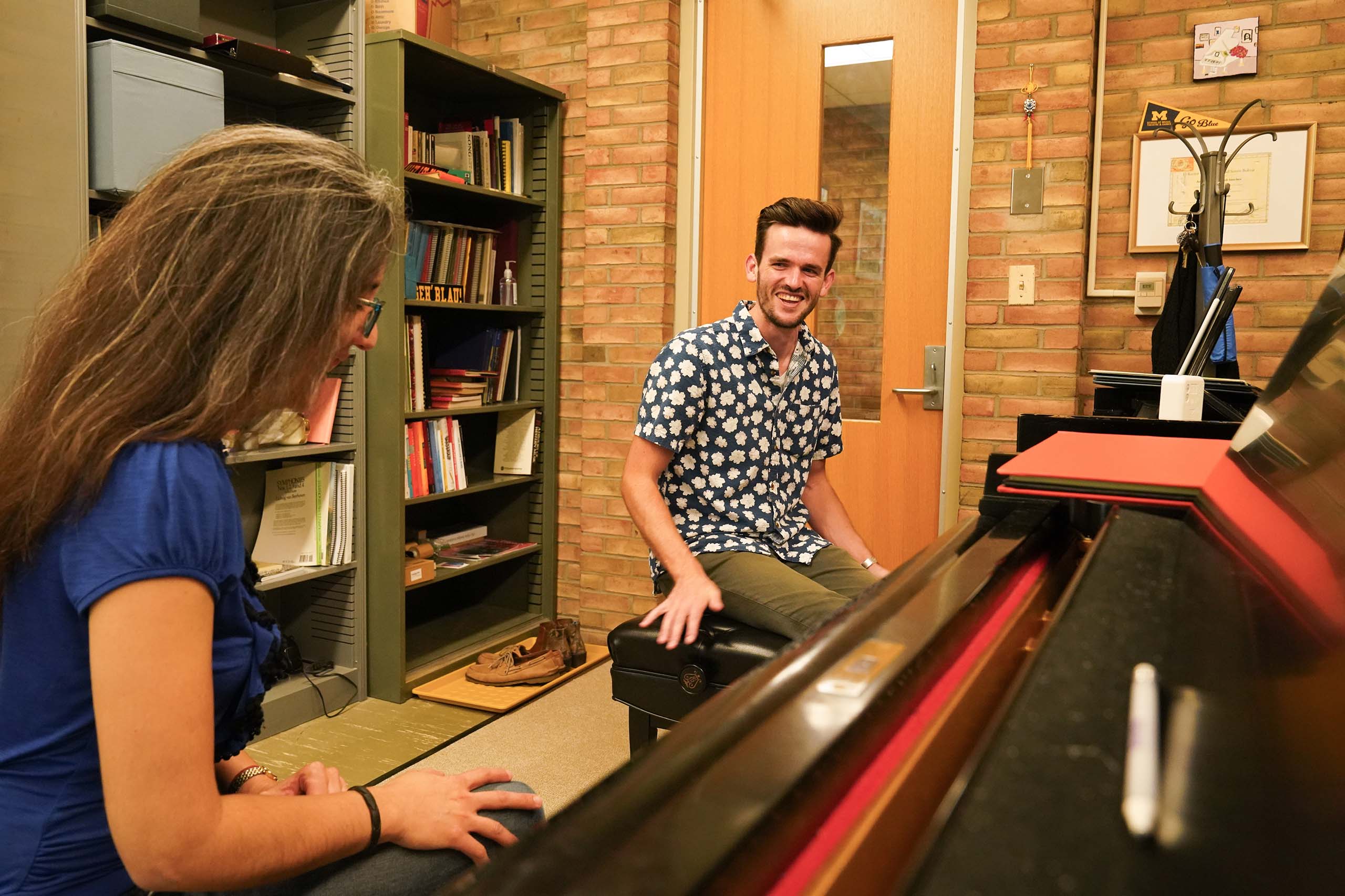 A student working with Professor Otamendi smiles, attired in a blue and white floral print shirt, with both seated at side-by-side pianos during a studio lesson.