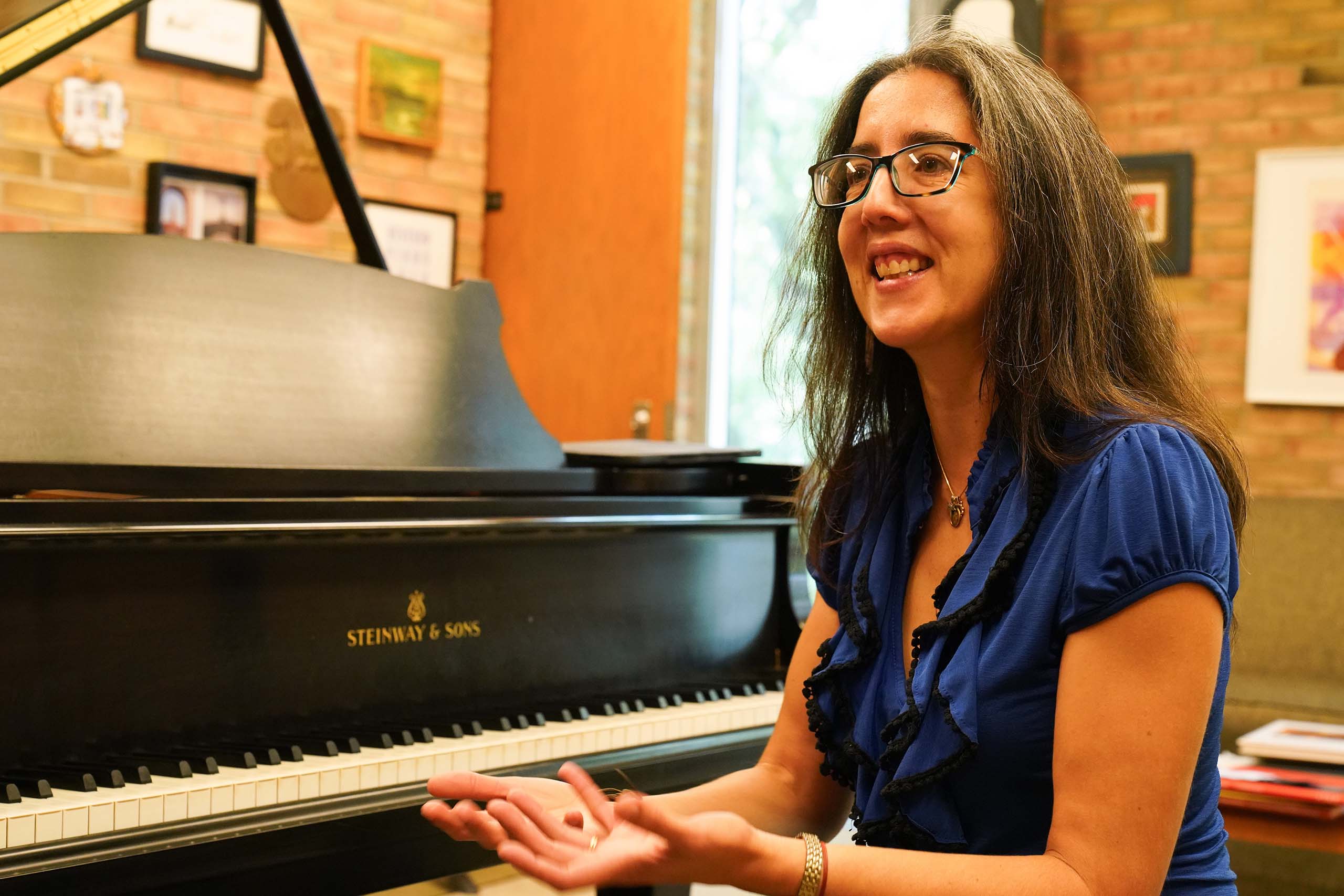 Professor Ana María Otamendi speaks seated at a piano during a studiolesson, wearing a blue blouse and glasses.