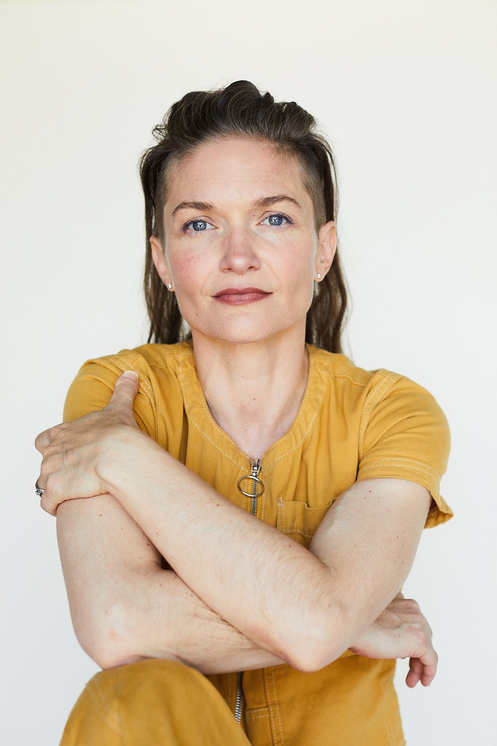 Studio portrait of Banning, attired in a marigold jumpsuit, seated with arms folded and a white backdrop.