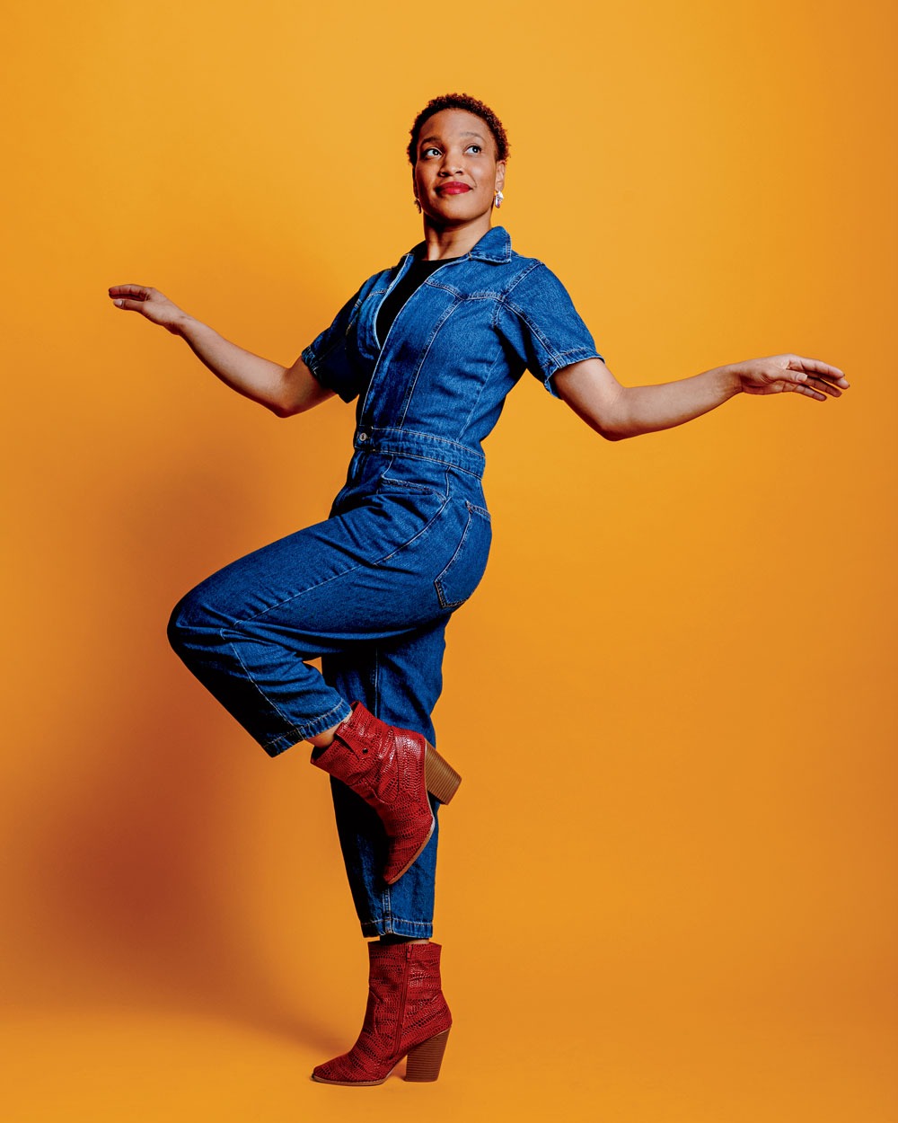 Studio portrait of Kia Smith, standing on one foot with arms held out, attired in a blue jean jumpsuit, with a yellow backdrop.