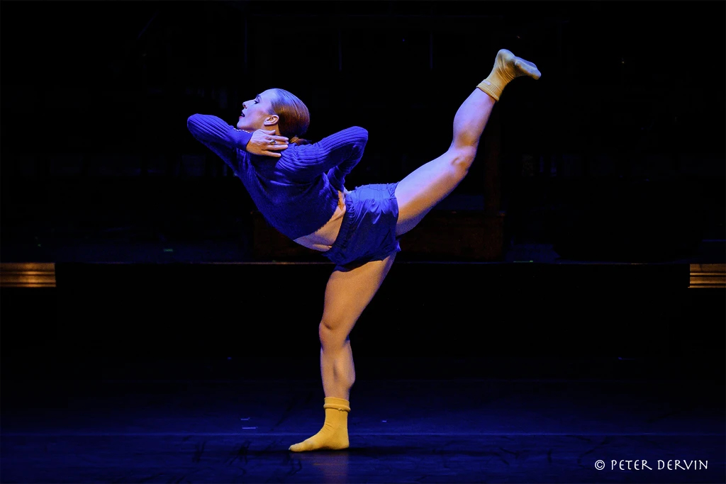 A dancer on a dark stage performs an arabesque with hands on the neck and back; attired in blue costume with yellow socks.