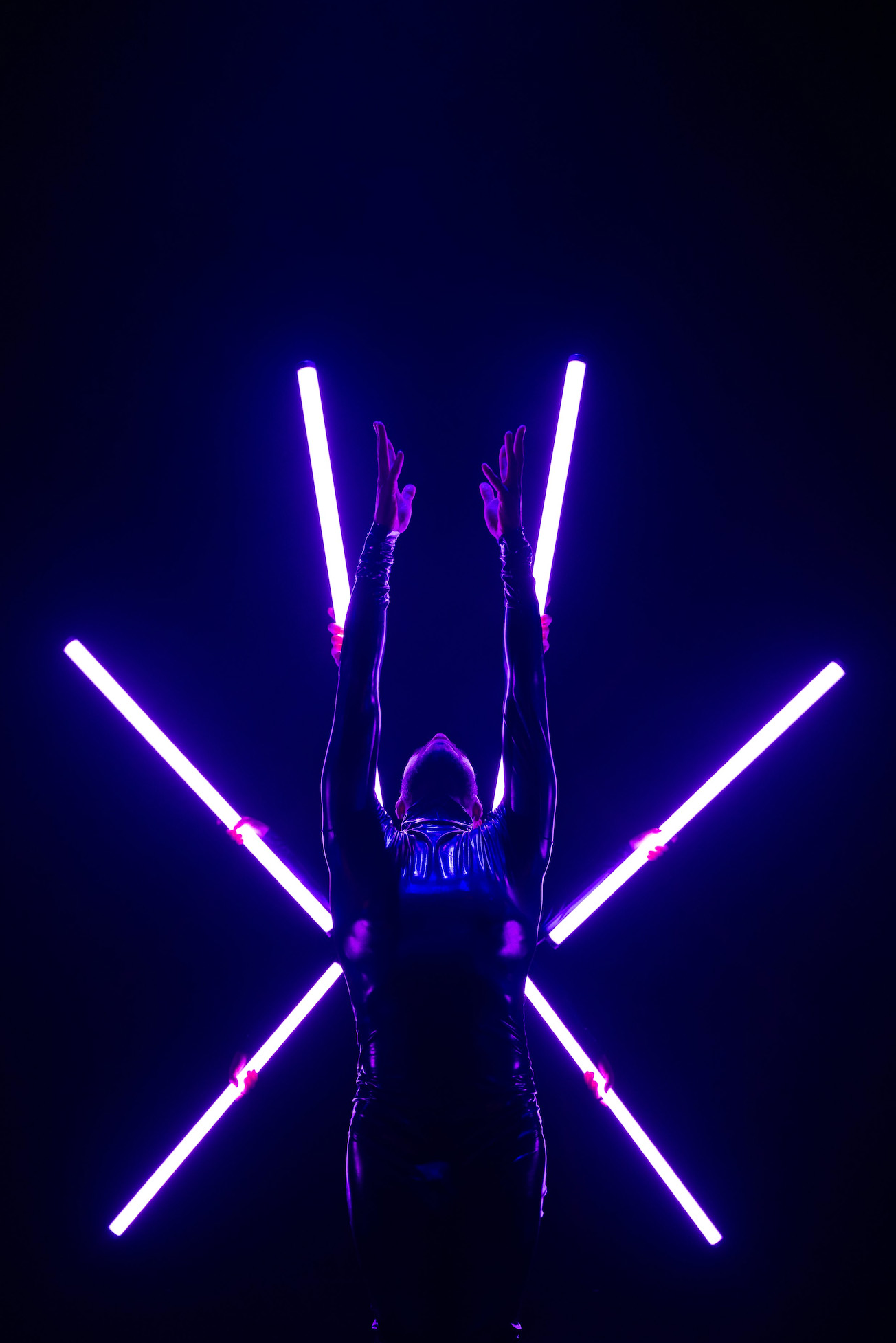 A dancer performs standing with arms and fingers pointed straight up, attired in dark costume with long white lights in the background.