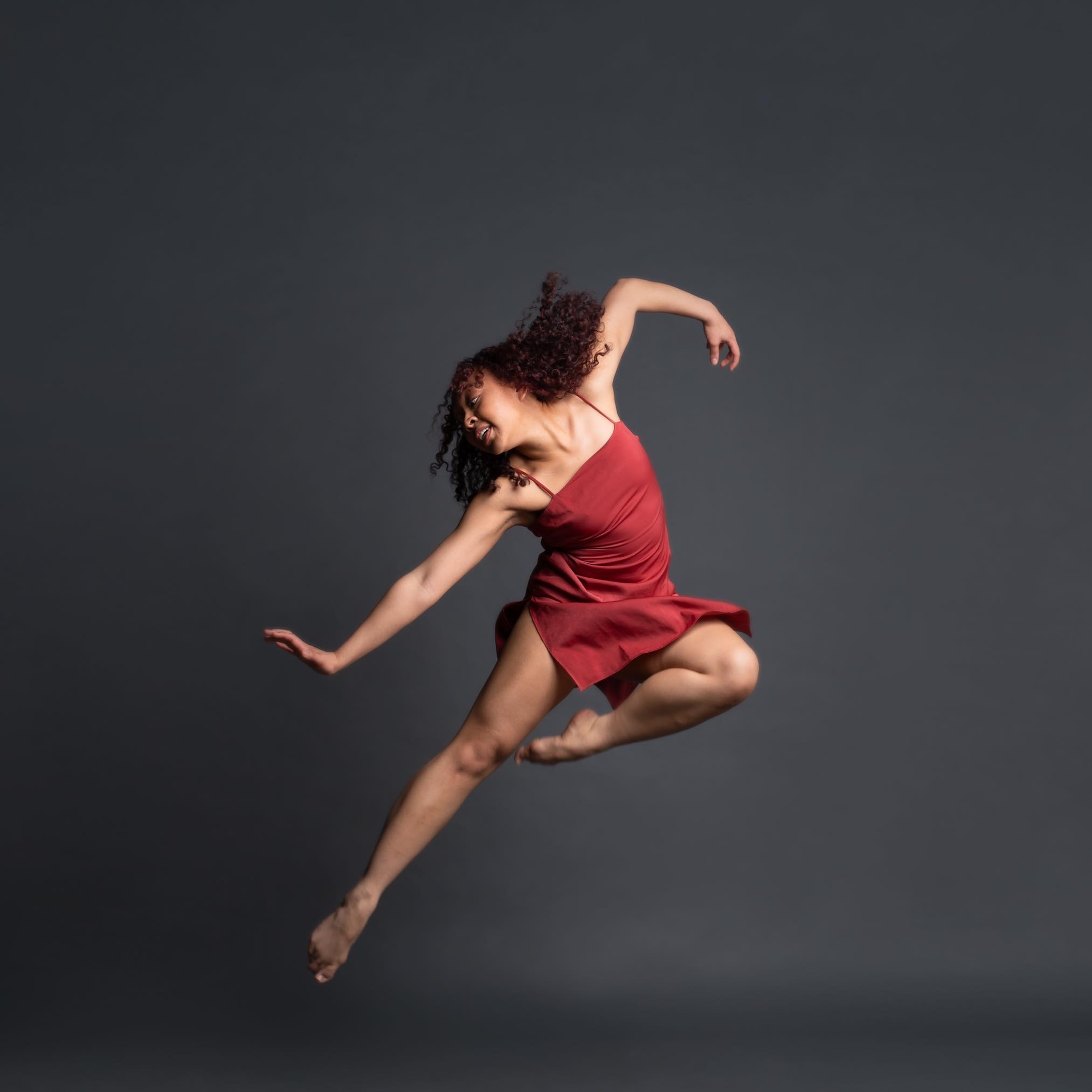 Studio portrait of a dancer leaping, with one knee and one elbow bent, attired in a red costume, with grey backdrop.