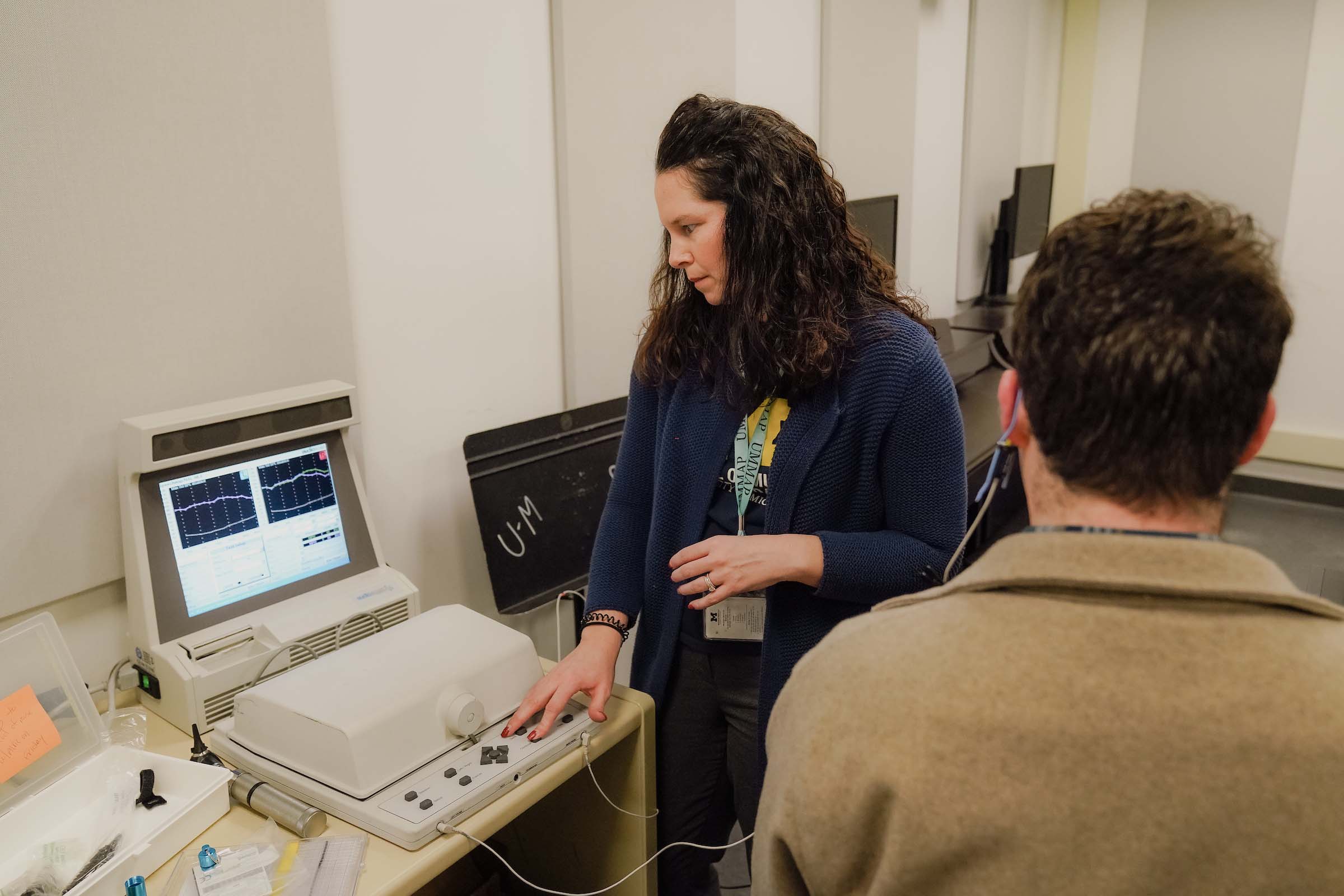 A clinician stands monitoring a table-top machine with a monitor, as a student sits next to the machine for an earplug fitting.