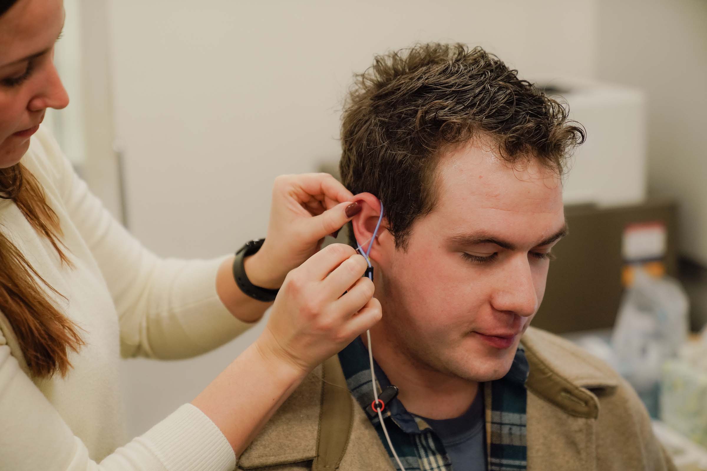 A clinician stands next to a seated student, holding two wire-like devices up on to their ear during an earplug fitting.