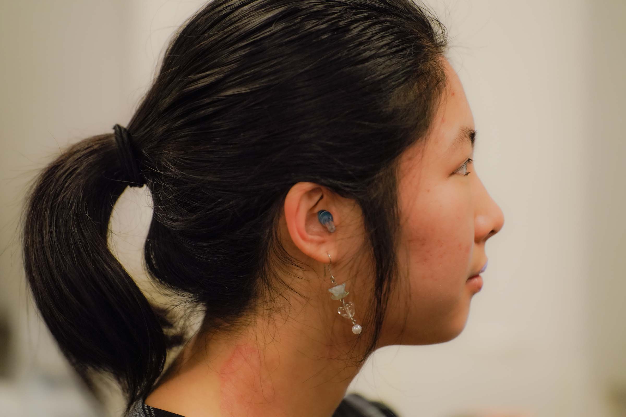 A student faces the right with an earplug visible in their ear, also wearing a dangling earring and hair pulled back in a ponytail.