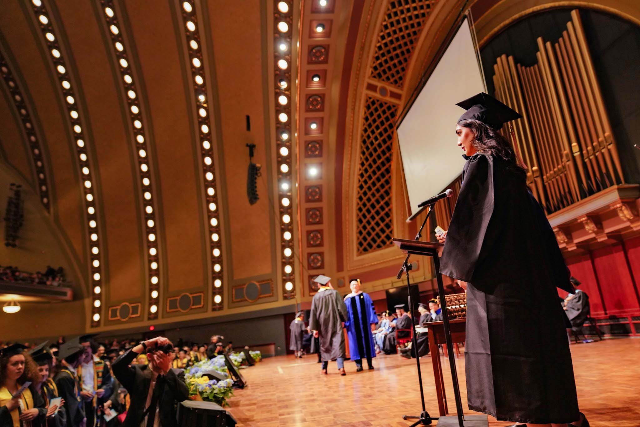 Graduating students attired in academic regalia cross the stage of Hill Auditorium to receive their diplomas, as the audience and platform party look on.