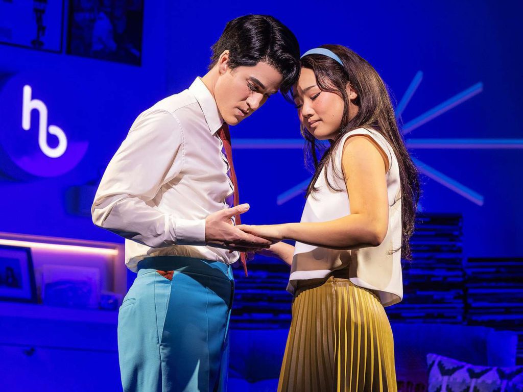During a performance of "Maybe Happy Ending," two actors perform standing on stage costumed in mid-20th century attire, with dark blue modern background.