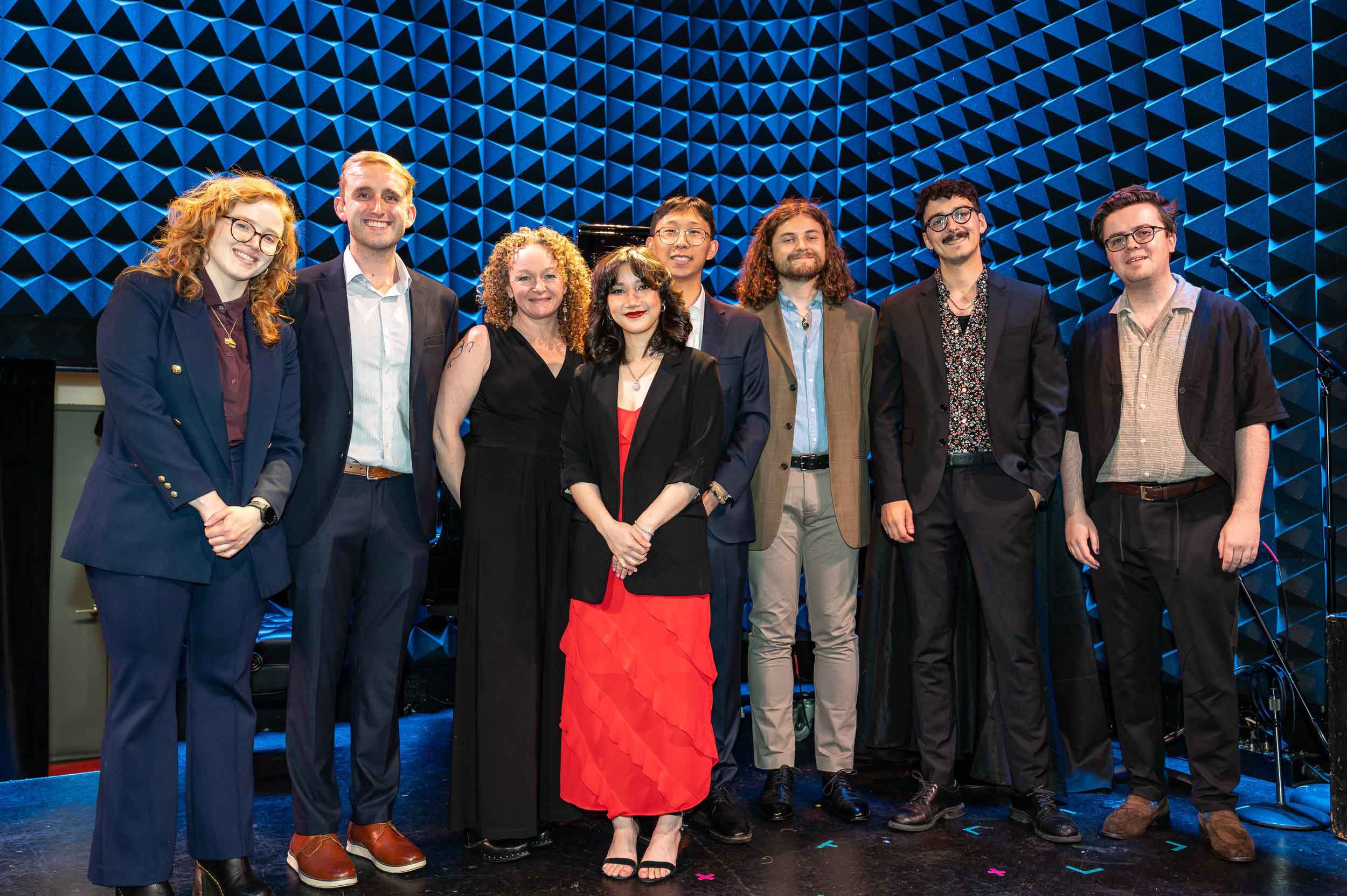 2025 BMI Composer Award winners Photo by Brian Berson Eight people pose together standing, wearing business casual attire, in a music studio space lined with dark blue sound treatments on the walls.