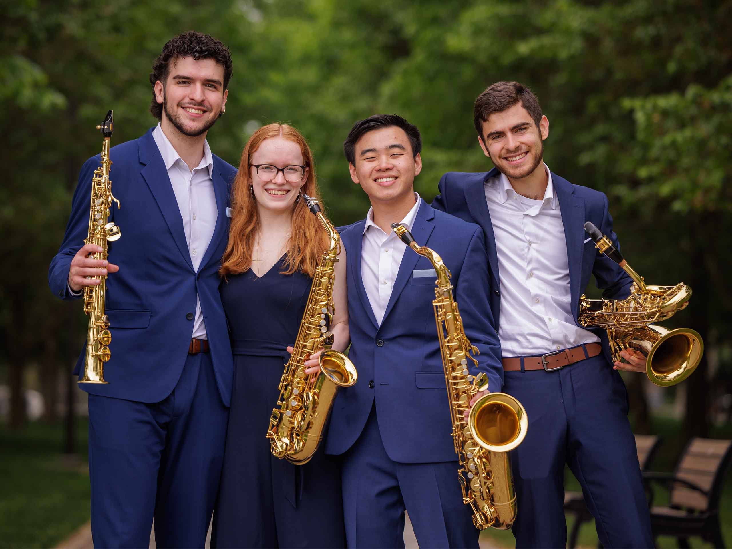 The four members of Cerus Quartet pose together standing outdoors in matching blue concert attire, holding their saxophones.