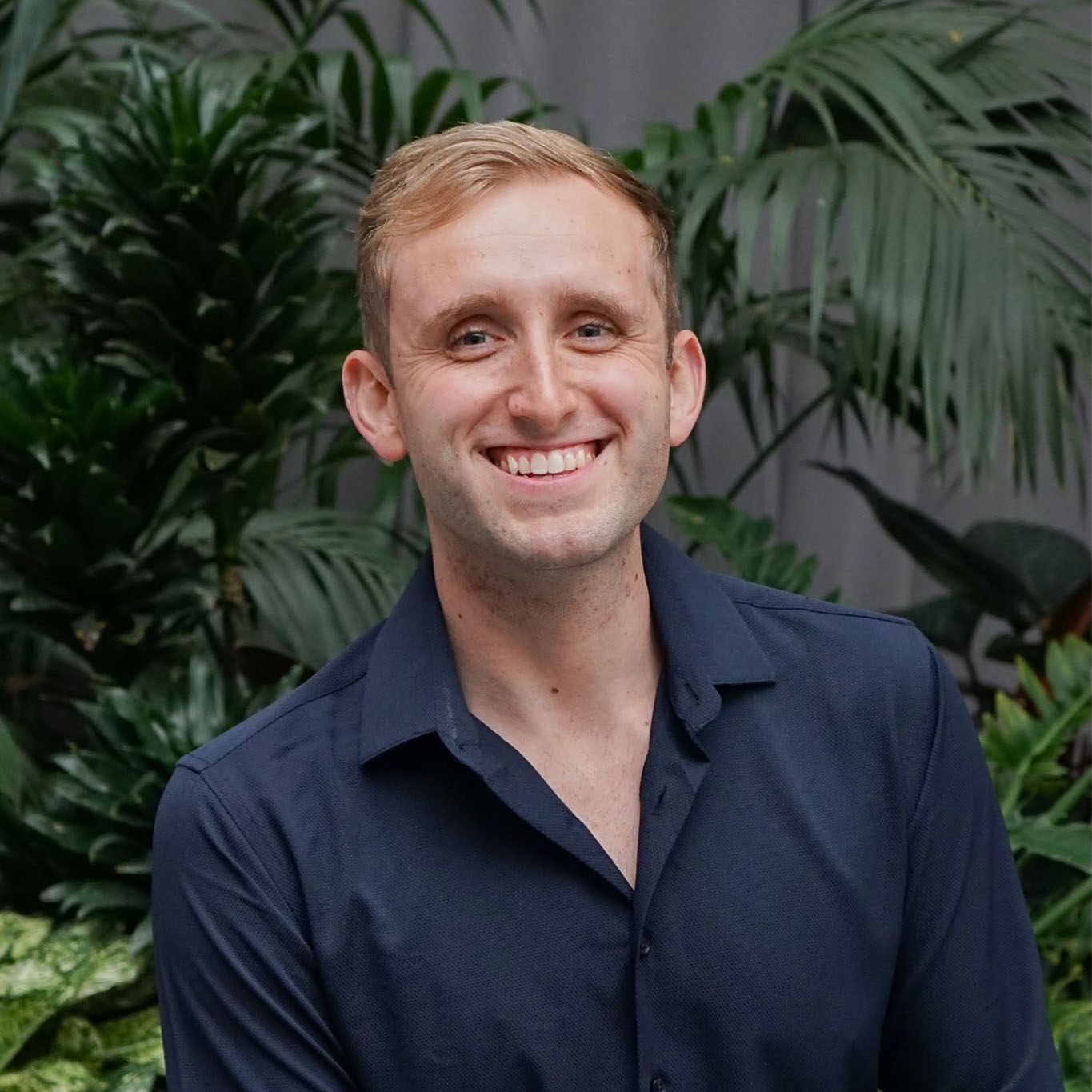 Cole Reyes 2025 BMI Composer Award Portrait of Cole Reyes wearing a dark blue collared shirt, with palm leaves and a dark grey wall in the background.