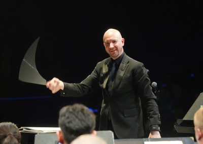 In performance on stage, Jason Fettig holds a conducting baton showing a trail from its quick movement; the heads of seated band members face him in the foreground.