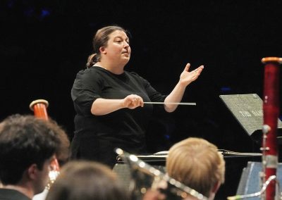 Attired in concert black, graduate assistant Kristina LaMarca conducts the band on stage, holding a baton with the other palm raised up.
