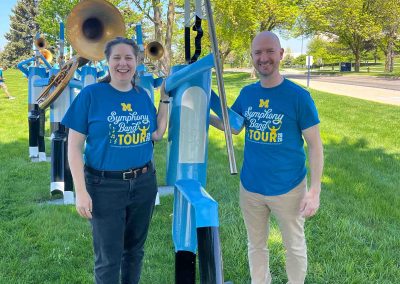 Assistant Dean Mica Harrison-Loosemore and Director of Bands Jason Fettig pose standing next to an outdoor marching band sculpture, both wearing blue Symphony Band Tour t-shirts.