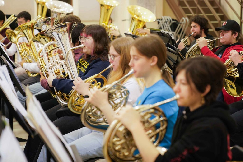Two rows of college and high school brass players seated in rehearsal, including French horns and euphoniums.
