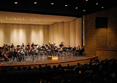 The Symphony Band performs on stage at Belleville High School auditorium, as Jason Fettig stands conducting with arms held high. The audience watches from a darkened house.