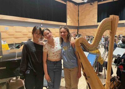 Three students pose standing in a band rehearsal, next to a harp and a piano.