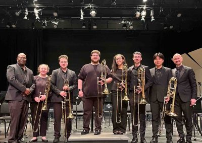 Six student trombone players stand on stage holding their instruments, posing with David Jackson and Jason Fettig.