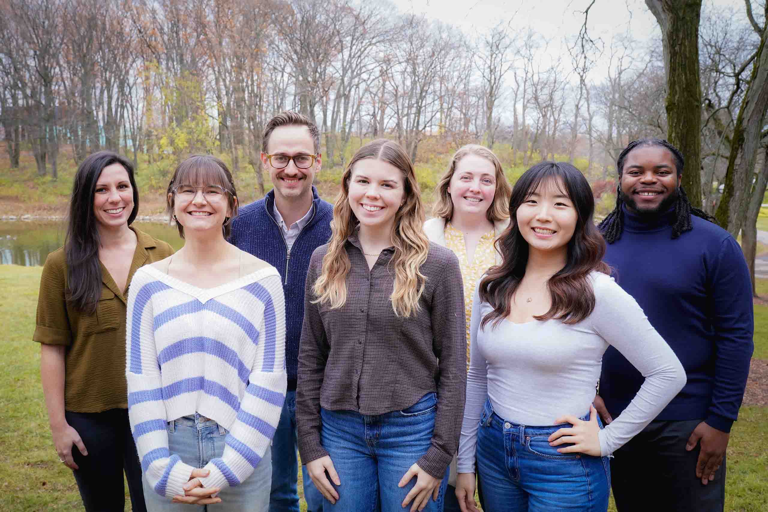 Seven people pose standing in two rows, outdoors with the SMTD Pond and late fall trees in the background.