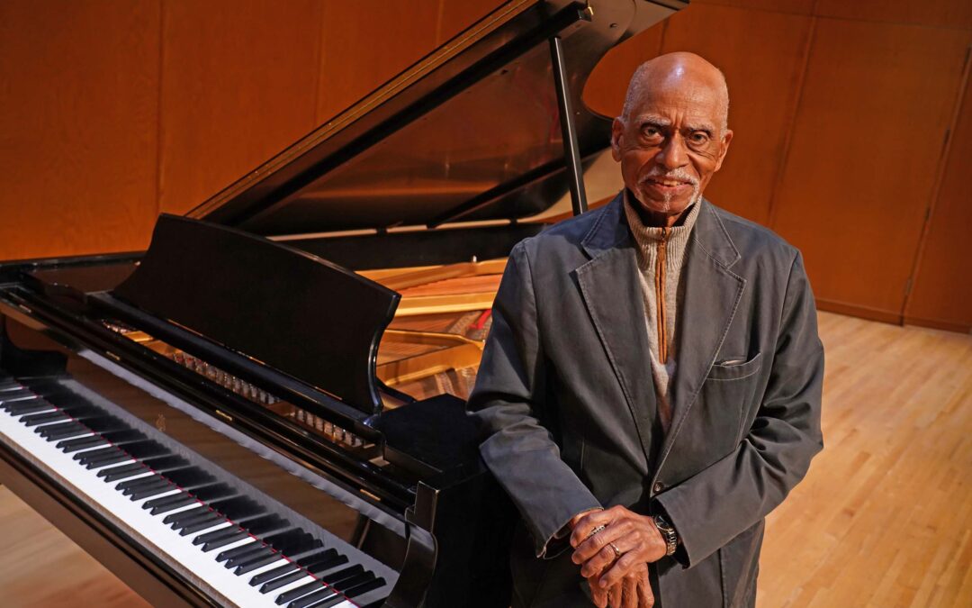 George Shirley poses on stage, attired in a casual suit coat, with his arm resting on a grand piano with its lid raised.