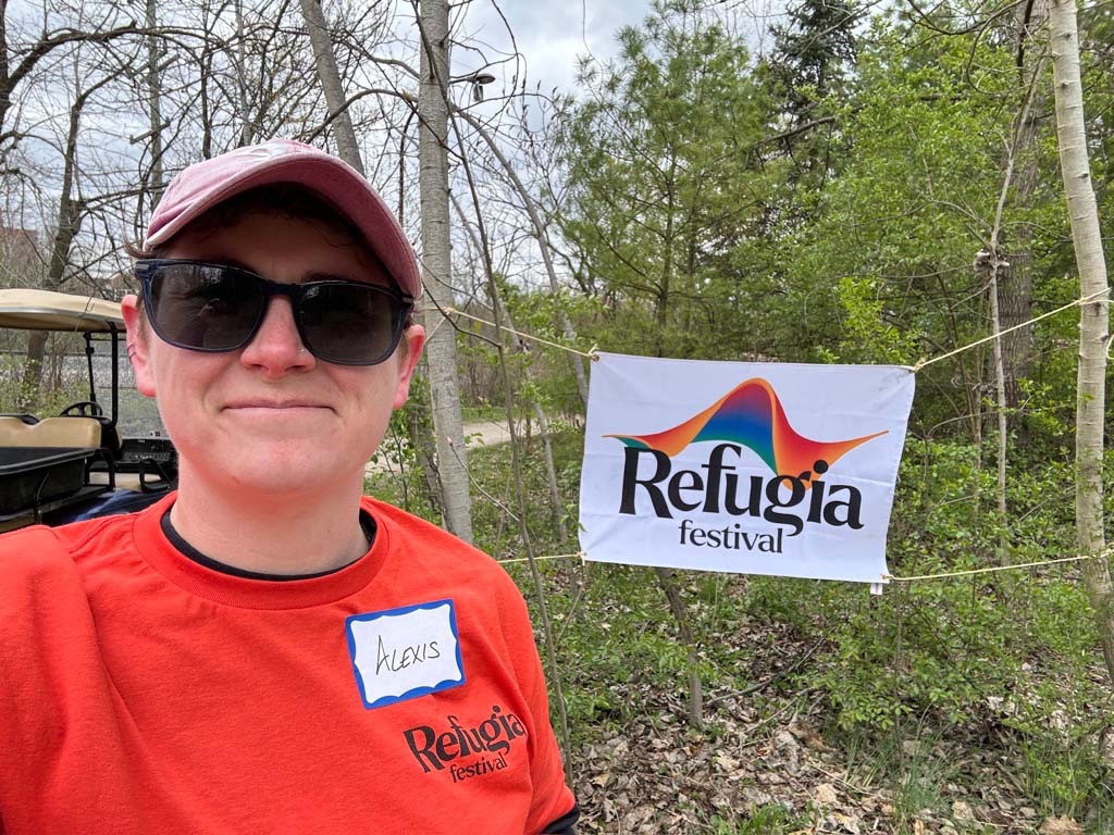 Alexis Lamb 2024 Refugia Festival Alexis Lamb selfie next to a Refugia Festival banner, outdoors in a wooded area.