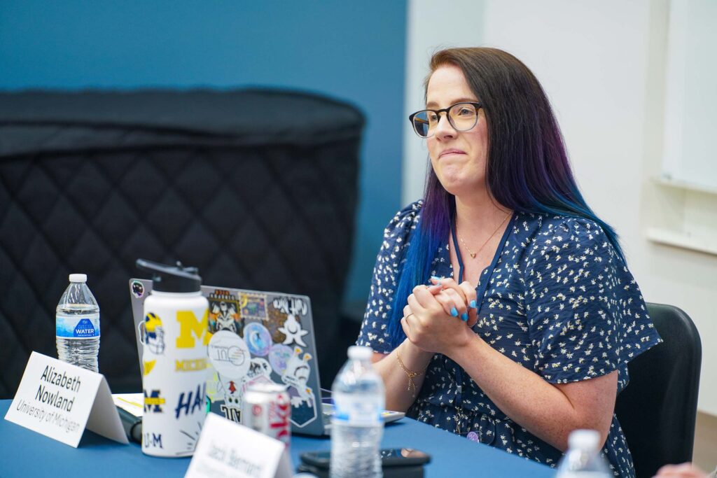 Nowland seated as a conference panelist, at a table with name plates, beverages, and computer.