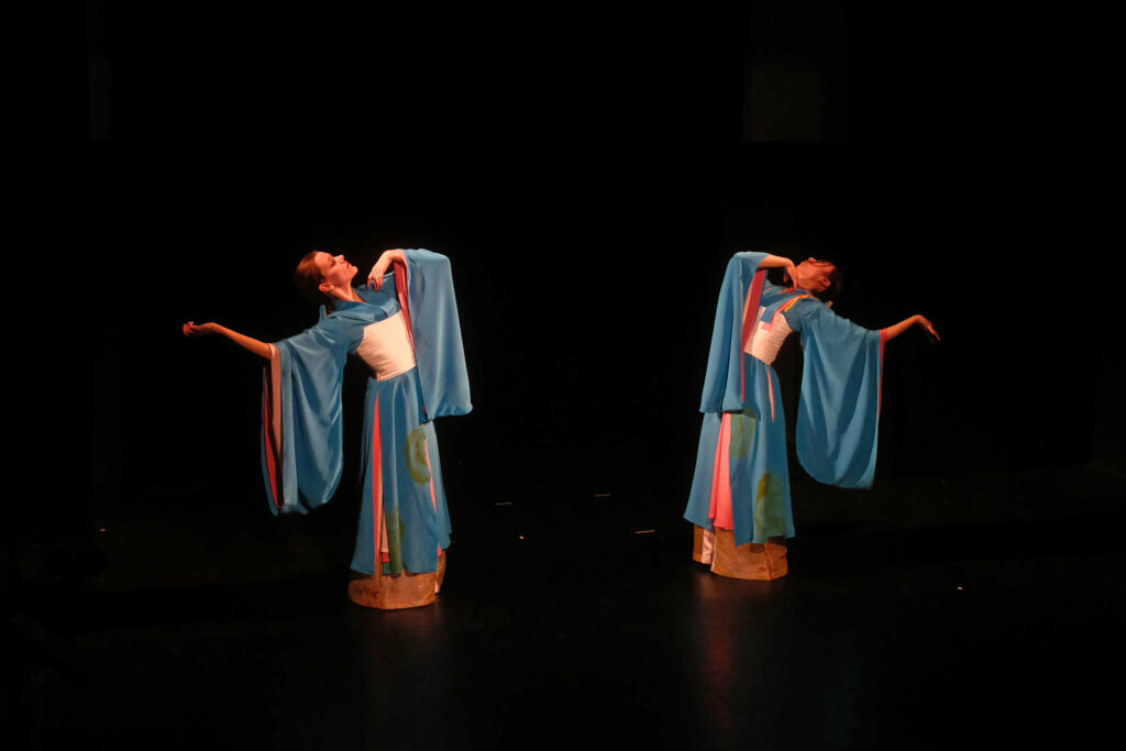 Two dancers costumed in matching blue and pink Chinese dress perform mirrored arm movements on a dark black stage.