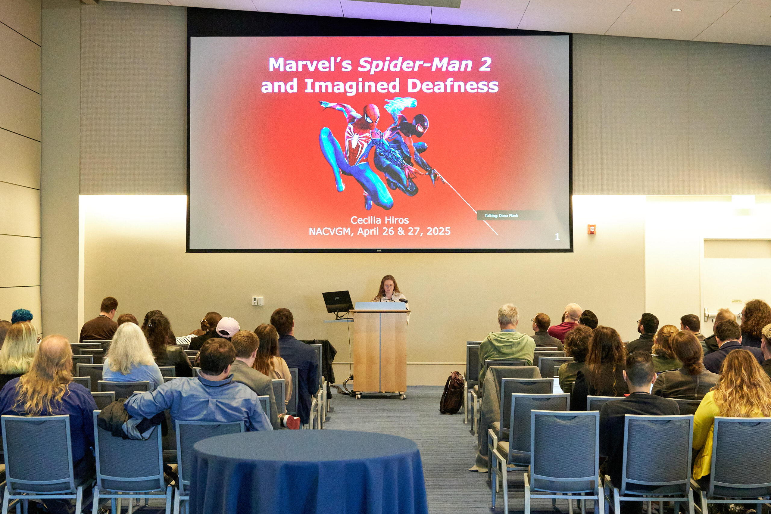 Cecilia Hiros presenting her research at the North American Conference for Video Game Music. Photo_ Sarah Pozderac-Chenevey Cecilia Hiros speaks at a podium below a large slide projection featuring Spider-Man, in a big conference space with seated audience.