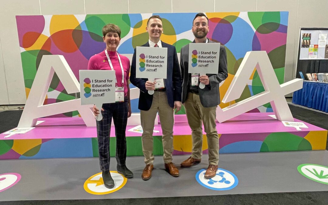 Three people pose with a colorful conference display, wearing nametags and holding up signs that say "I stand for education research."