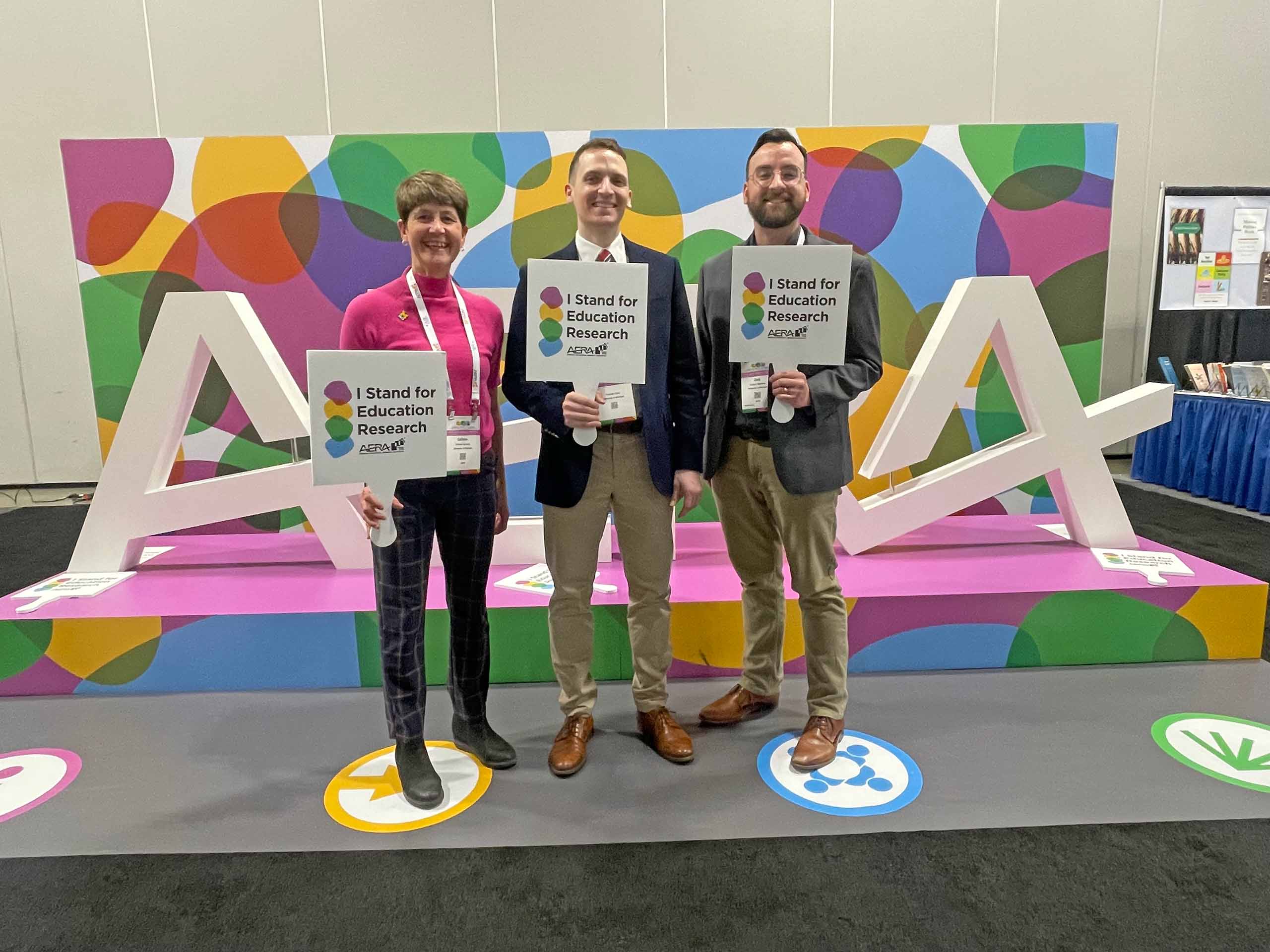 Three people pose with a colorful conference display, wearing nametags and holding up signs that say "I stand for education research."