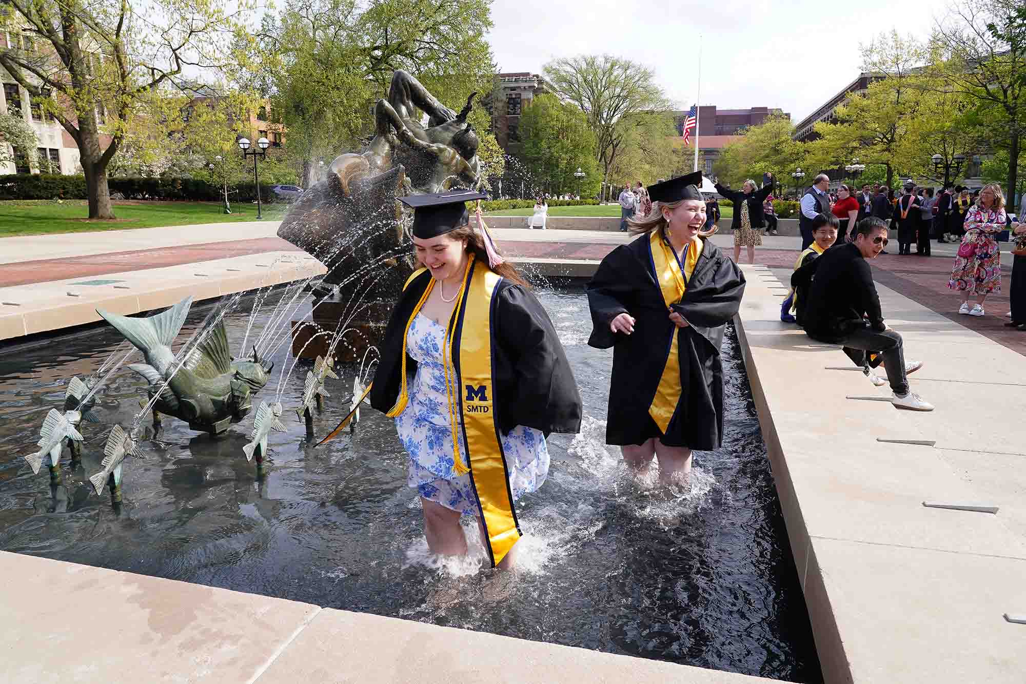 Commencement SMTD students celebrate May 2025 Two graduating students in regalia walk through the fountain next to Hill Auditorium after the SMTD Commencement.