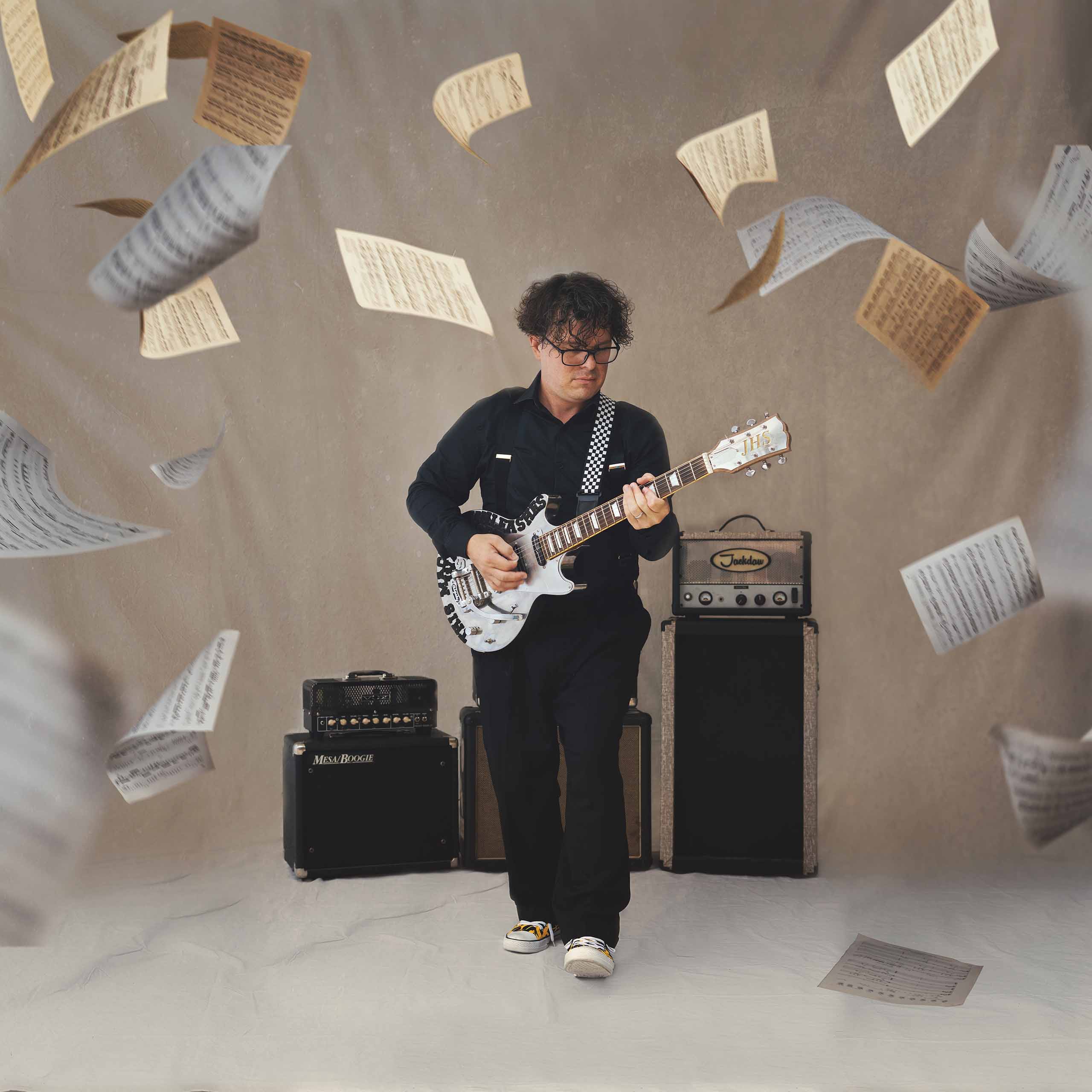 In a photo studio, D.J. Sparr plays electric guitar standing in front of amps,  as sheet music flies around the space.
