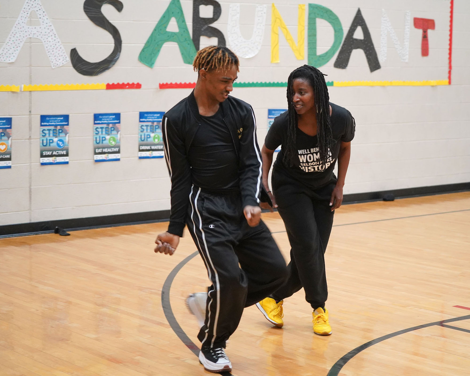 A dance instructor works with a student on moves in a school gymnasium.