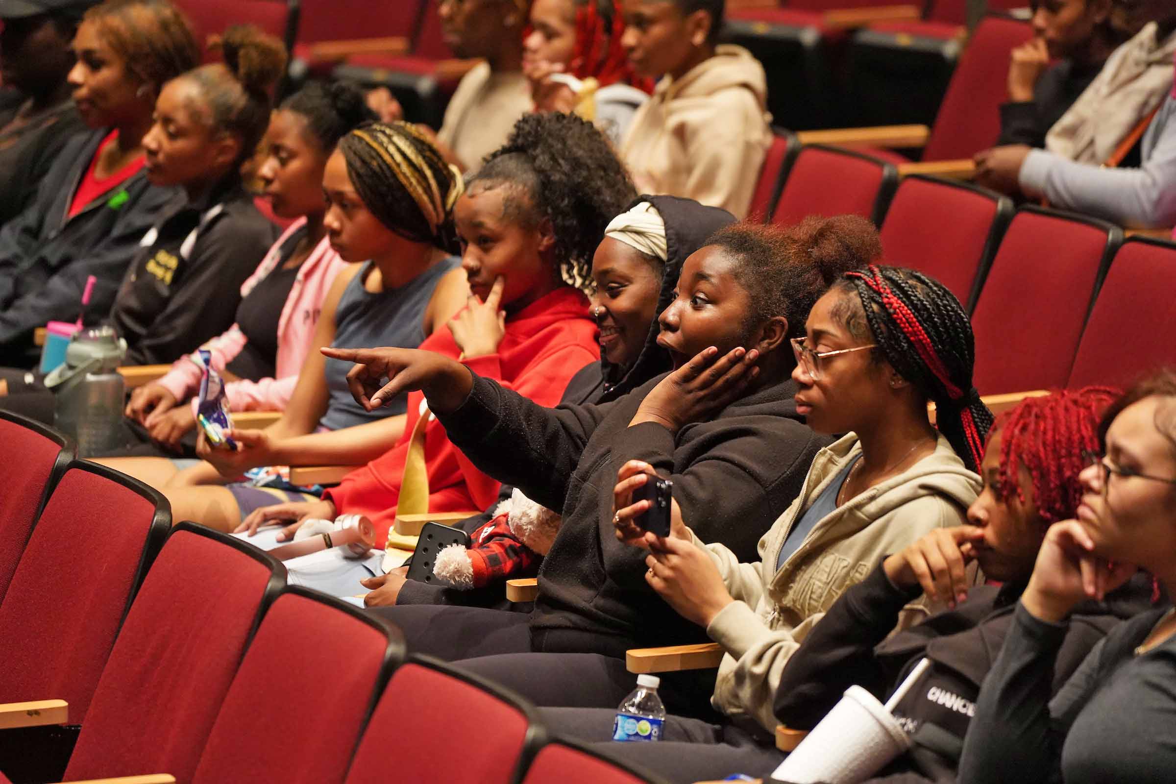 A row of students seated in a theatre react to something on stage.