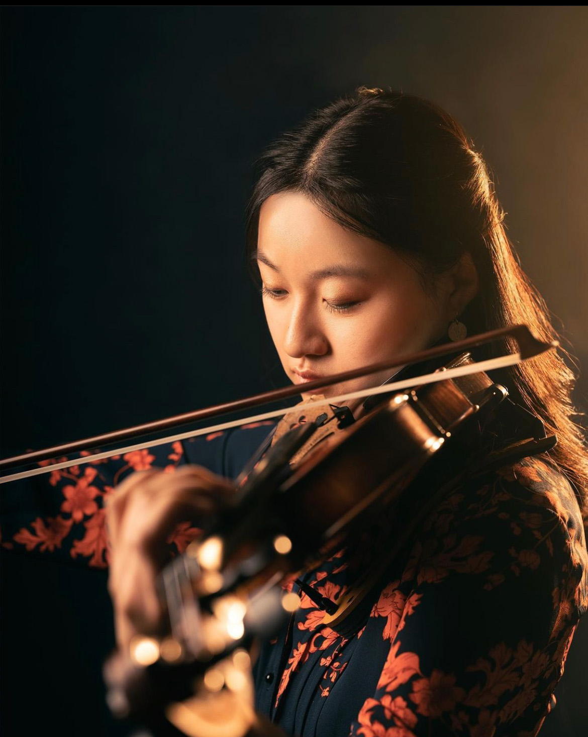 Ellen Hayashi Studio portrait of Ellen Hayashi playing violin, with black background and warm gold lighting.