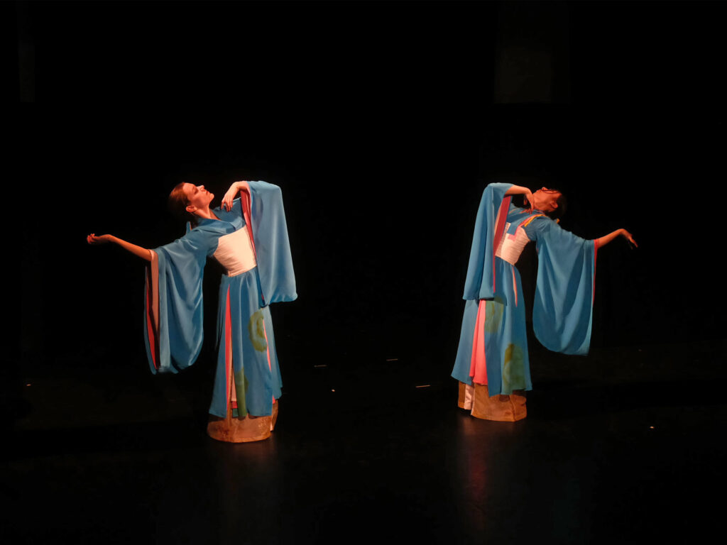 Two dancers costumed in matching blue and pink Chinese dress perform mirrored arm movements on a dark black stage.