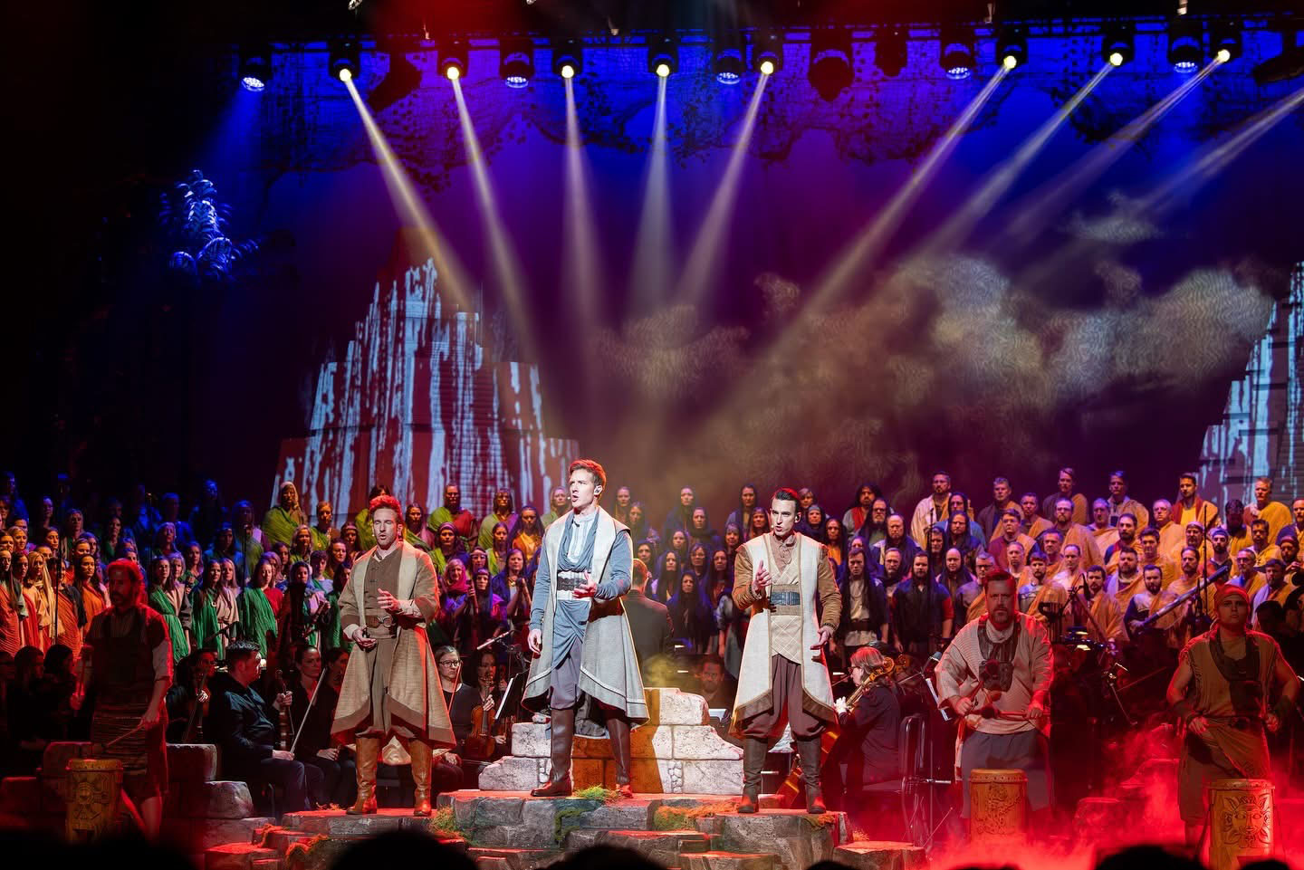 A performance of featured musicians, with background choir filling the stage; with dramatic lighting of blue, red, and projected natural textures.
