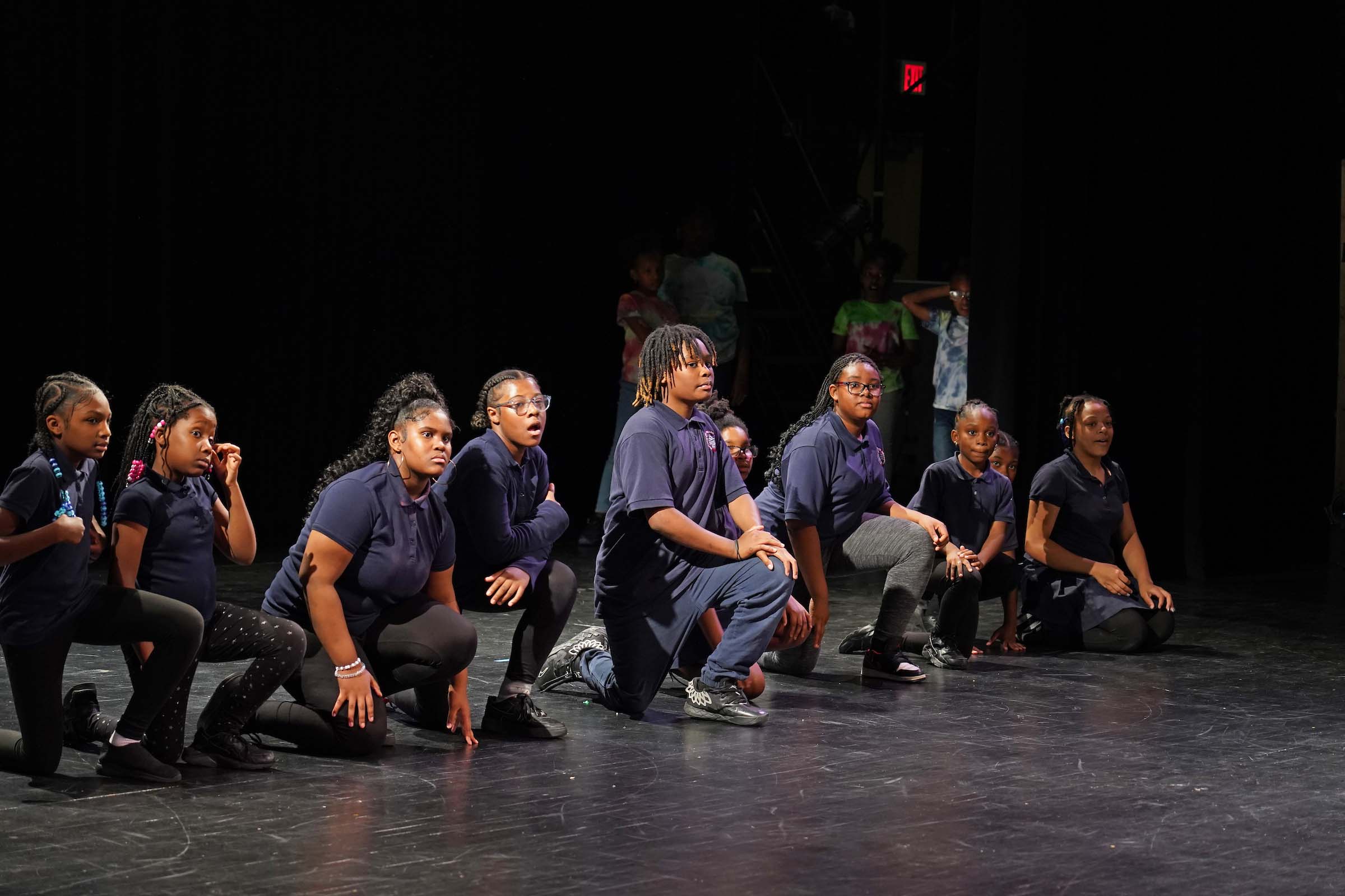Young students perform in marching costumes on a stage, all bent onto one knee.