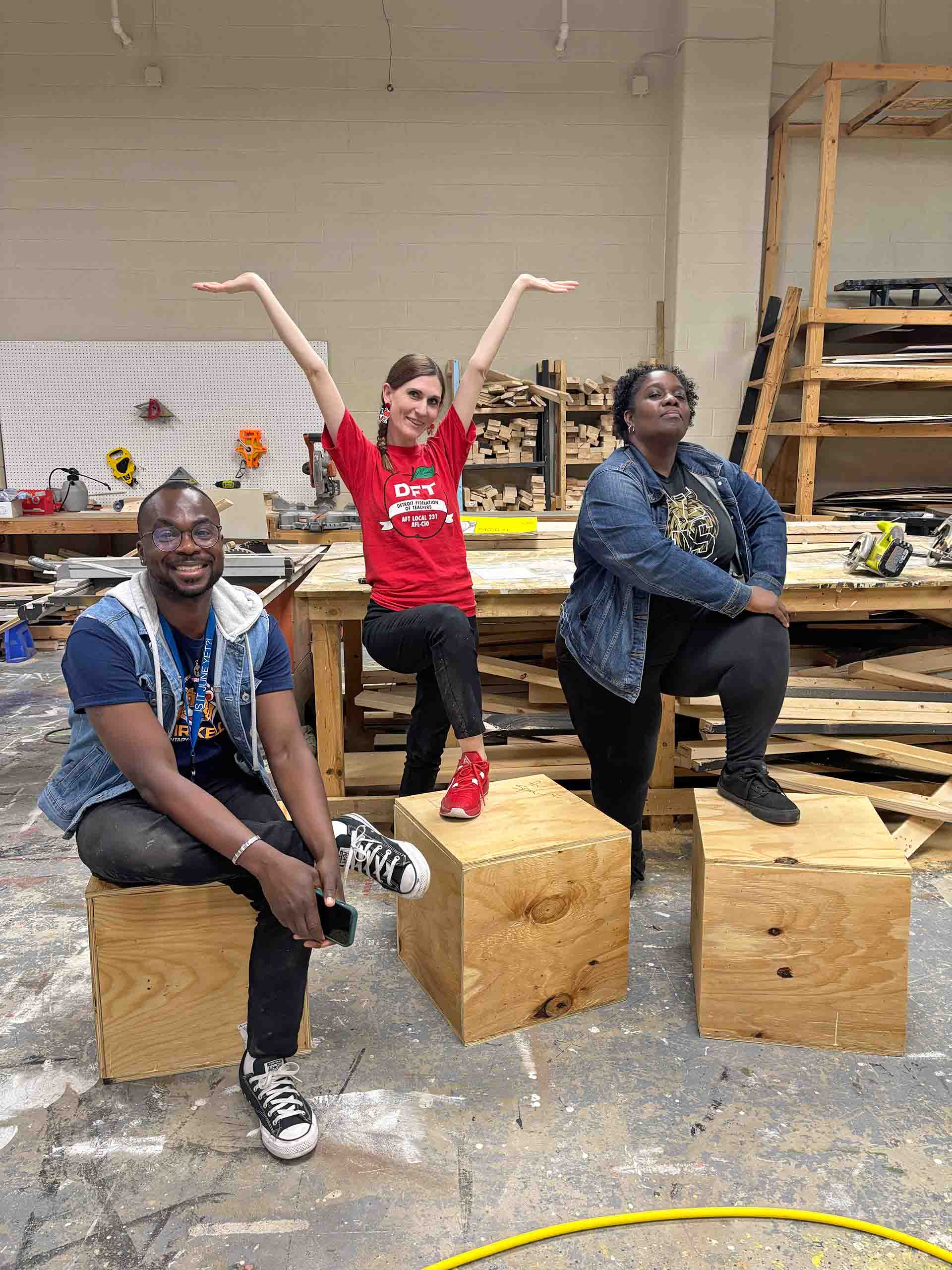 Three teachers pose on crates in a theatrical scenery shop.