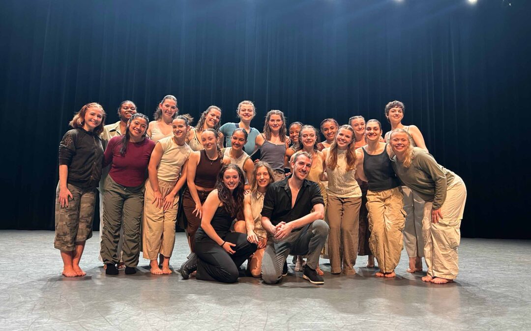 About 21 dancers pose together as a group, all attired in neutral, natural fabrics, in a studio space under stage lighting.
