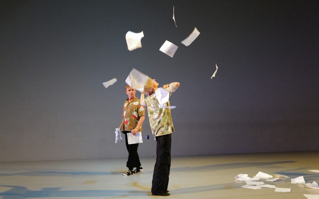Two actors perform on a sparse stage, with papers thrown in the air and on the ground; costumed in matching black pants and flower shirts.
