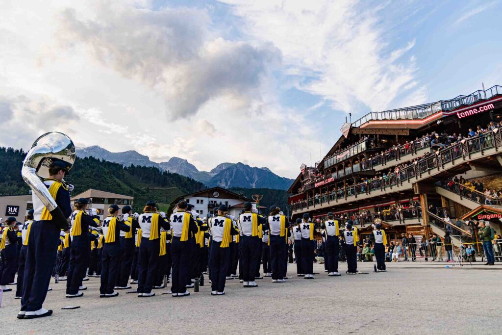 The Michigan Banding Band performs in formation outdoors, facing a crowd on a terraced building and high mountains in the distance.