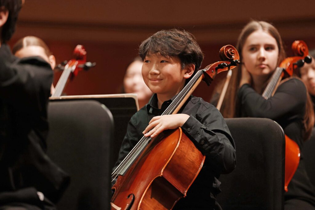 A youth cellist smiles while seated in the cello section at a performance.