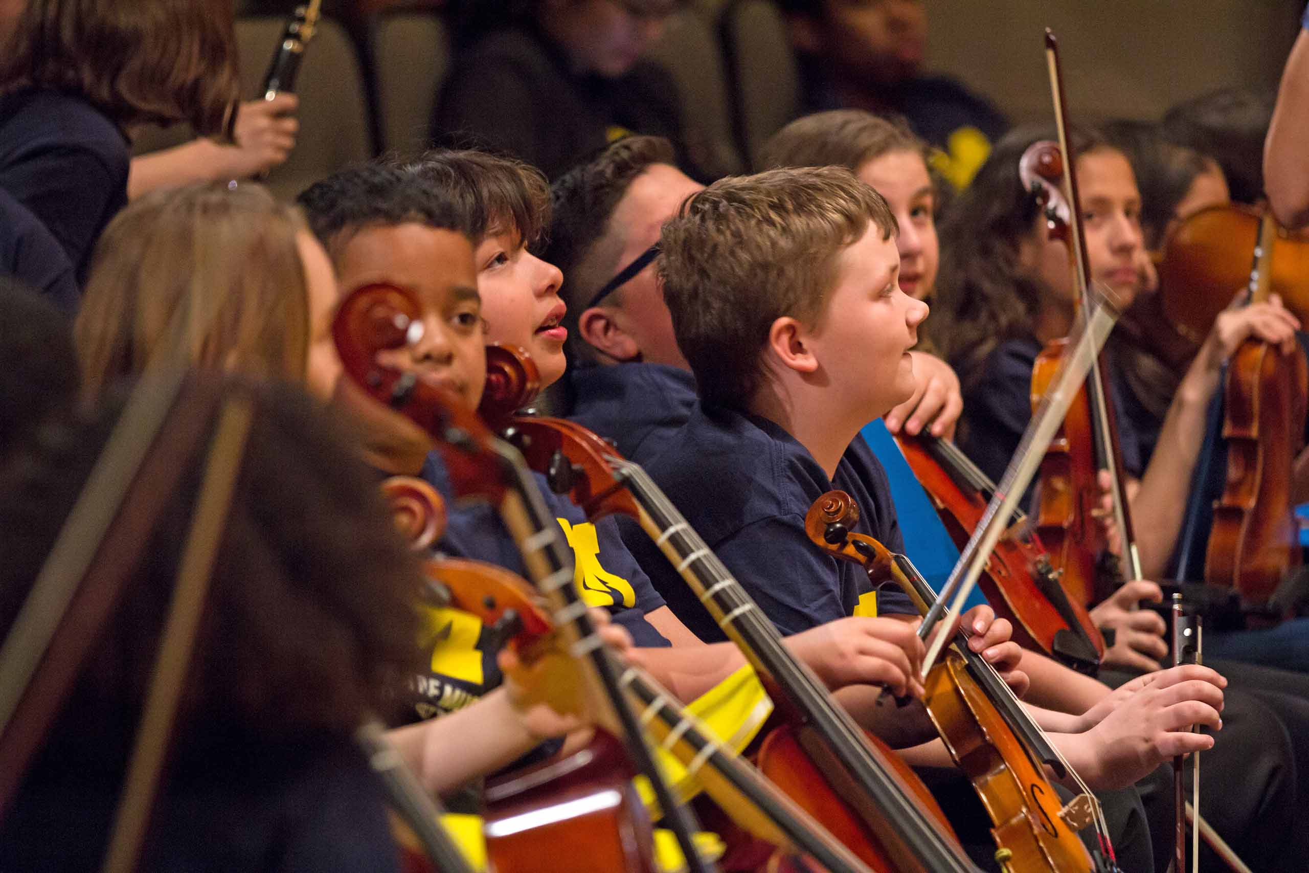 A row of elementary students seated holding string instruments, all wearing blue SMTD t-shirts.