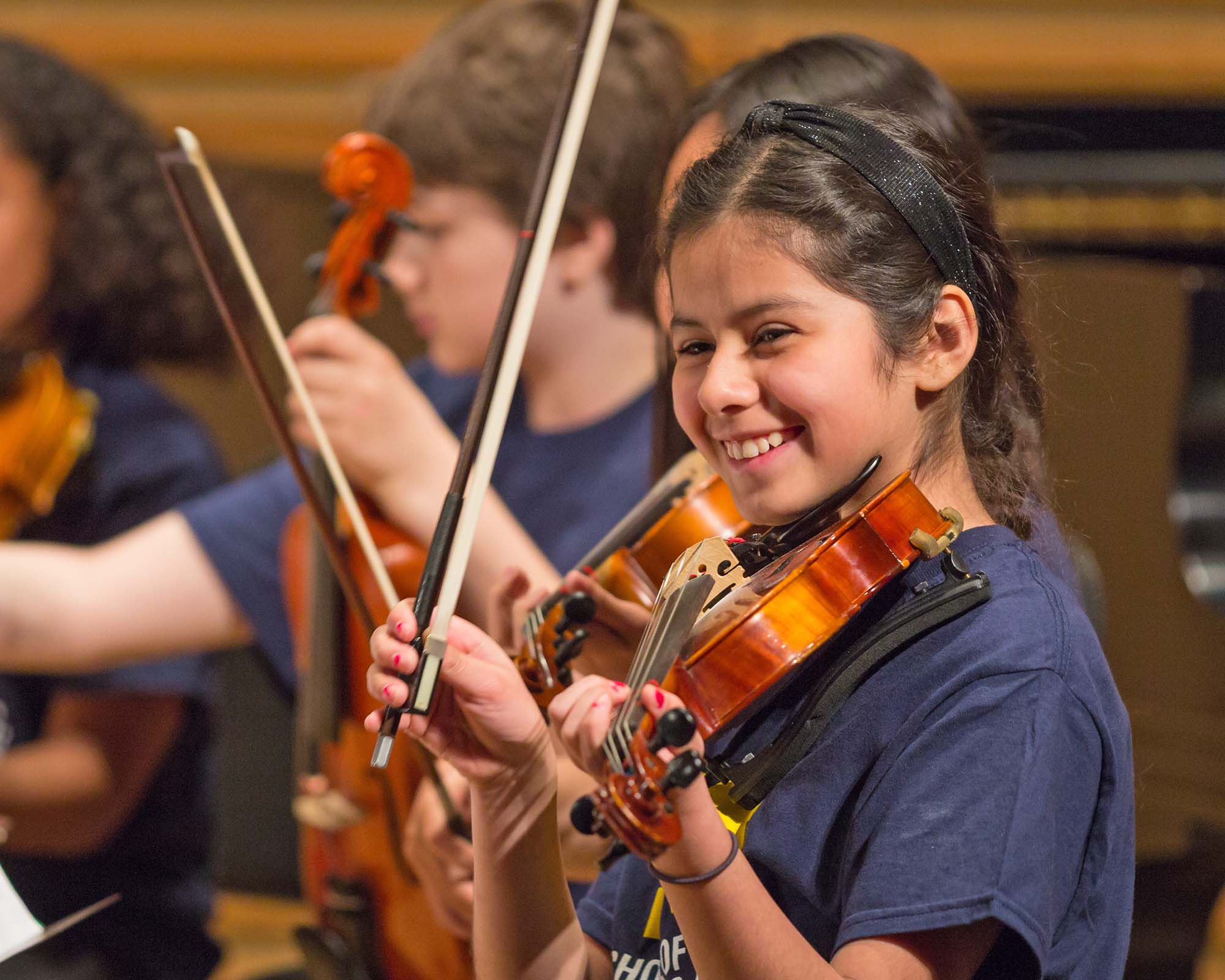 An elementary student smiles and holds their violin while seated with peers in a performance.