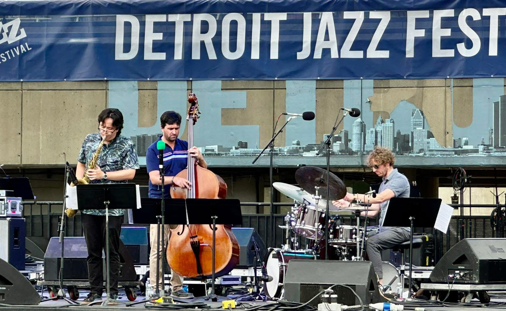 On a Detroit Jazz Festival outdoor stage, the trio of saxophone, bass, and drums performs.