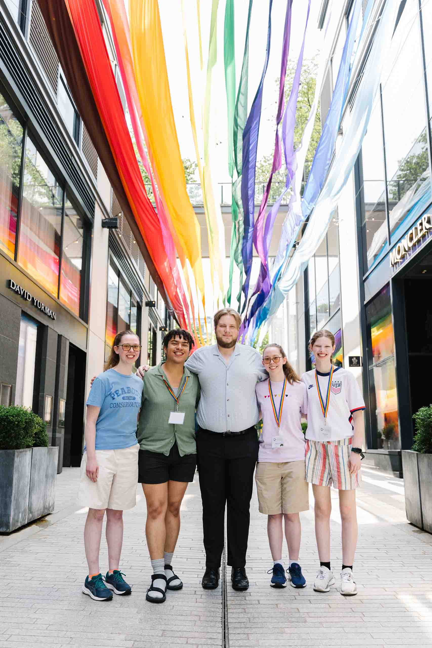 OUT Ensemble in DC Photo Da Ping Luo Five students pose together standing in casual attire, with rainbow-colored banners suspended above.