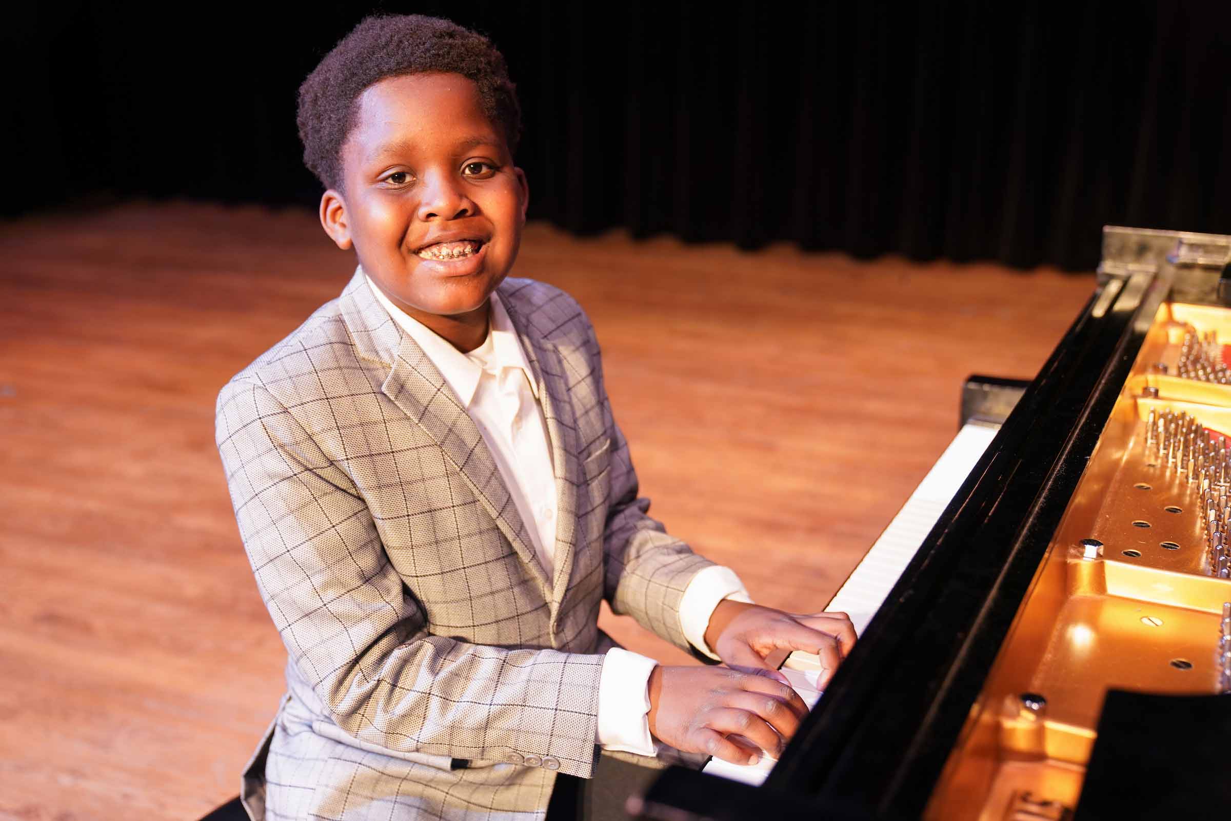 A young student smiles posing in position to play a piano, wearing a grey suit coat.